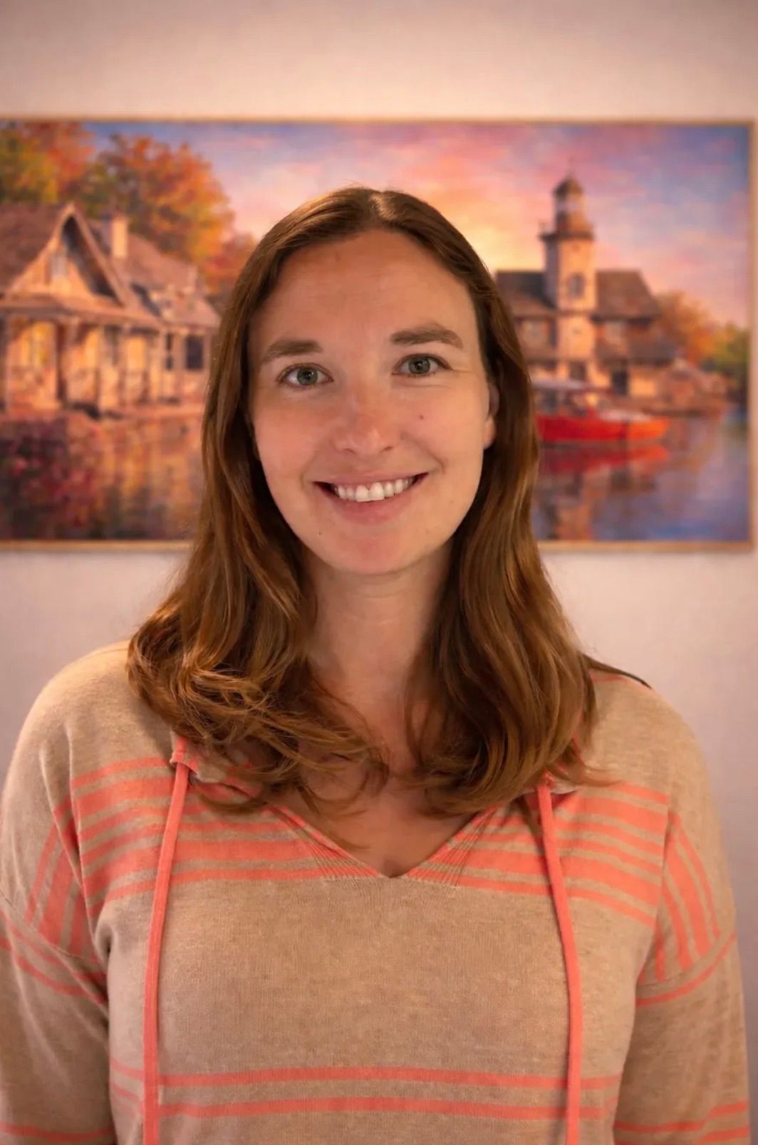 Heather Catherine, uma mulher sorridente de cabelo comprido castanho-claro, com uma camisola bege de riscas cor de coral, em frente a um quadro de uma aldeia à beira-rio ao pôr do sol.