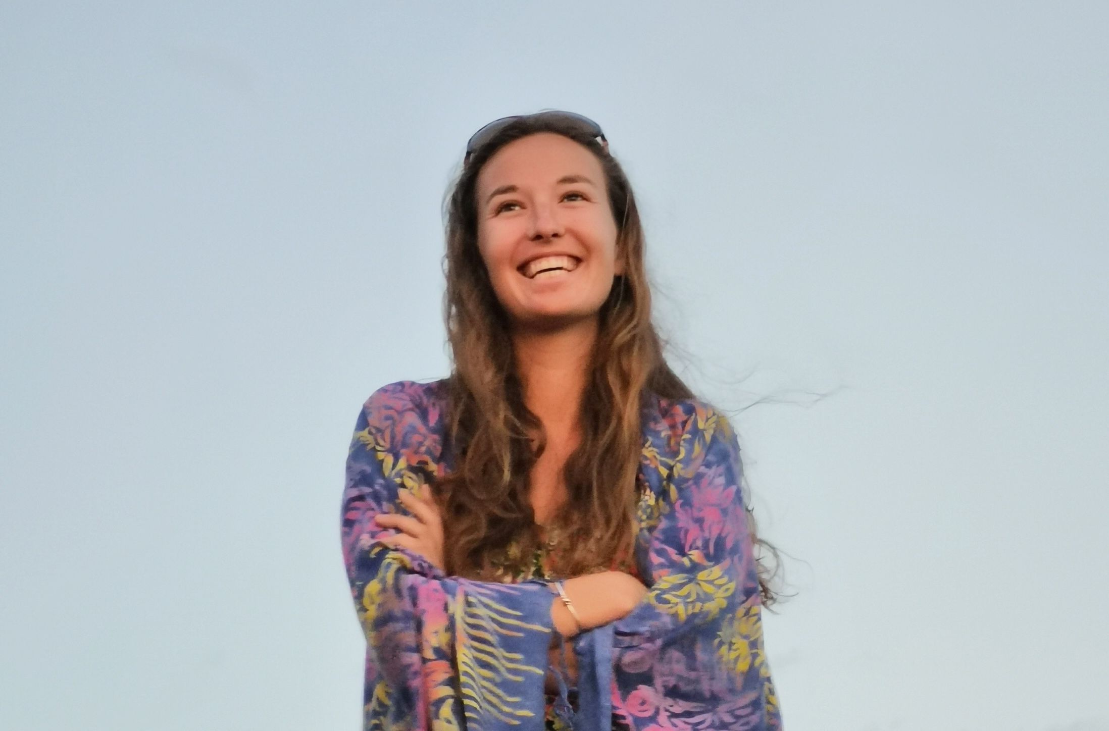 Heather Catherine a rir ao ar livre contra um céu azul claro, de braços cruzados, com óculos de sol na cabeça e uma blusa florida em tons de roxo e amarelo.